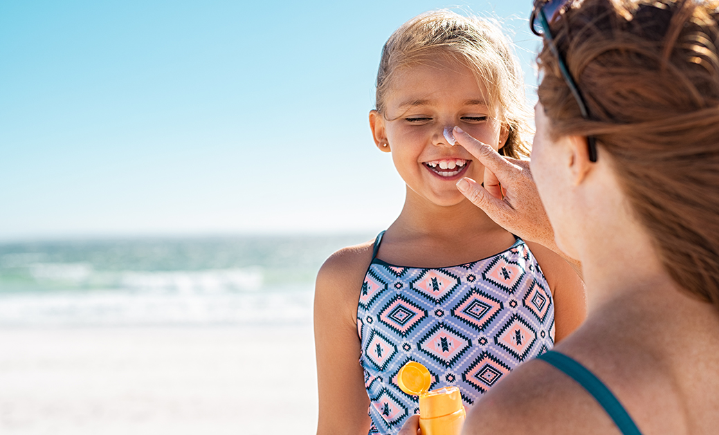 protector solar infantil, imagen de una mujer aplicando crema solar a una niña en la playa